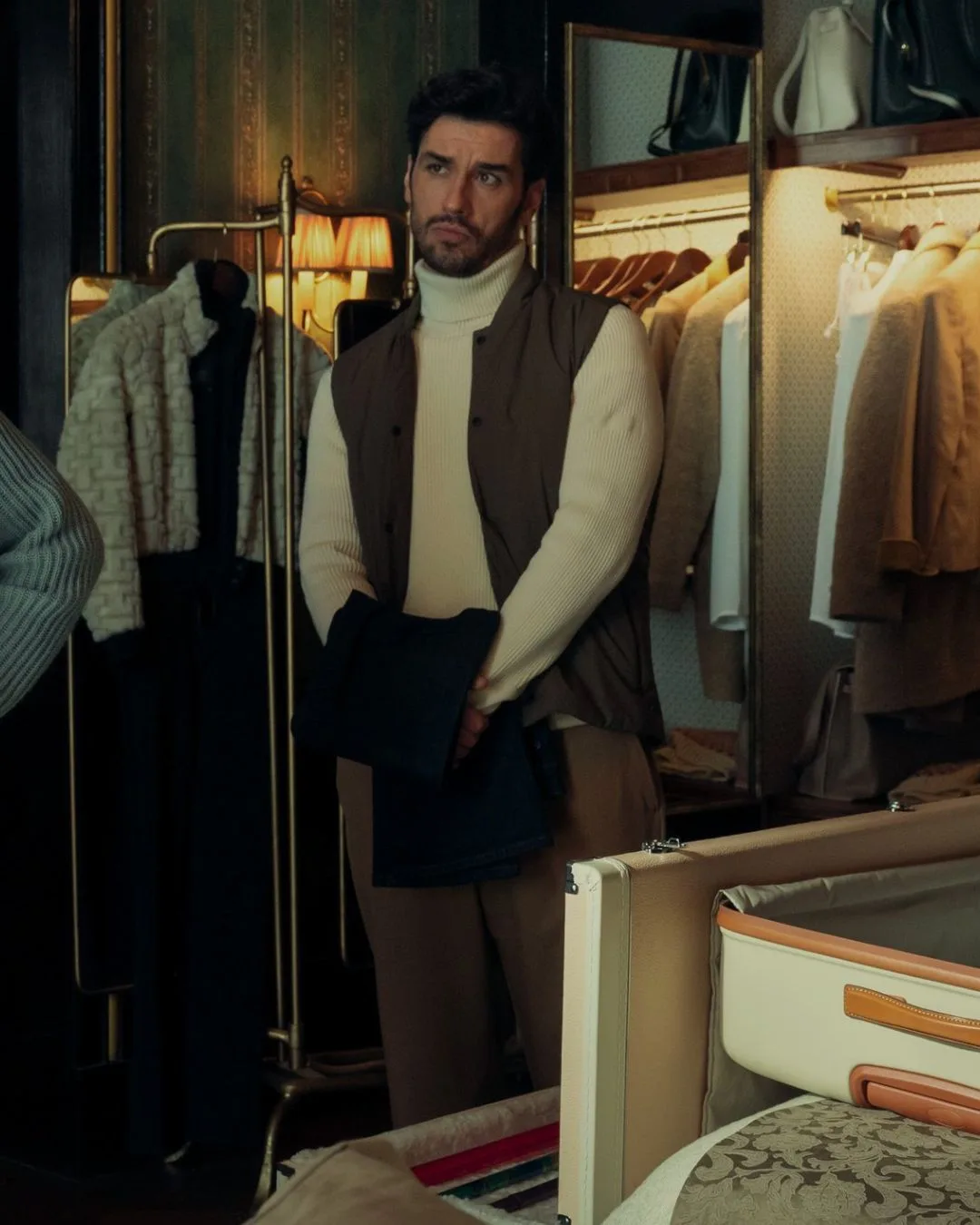 A man wearing a white turtleneck top and taupe vest staring seriously. 