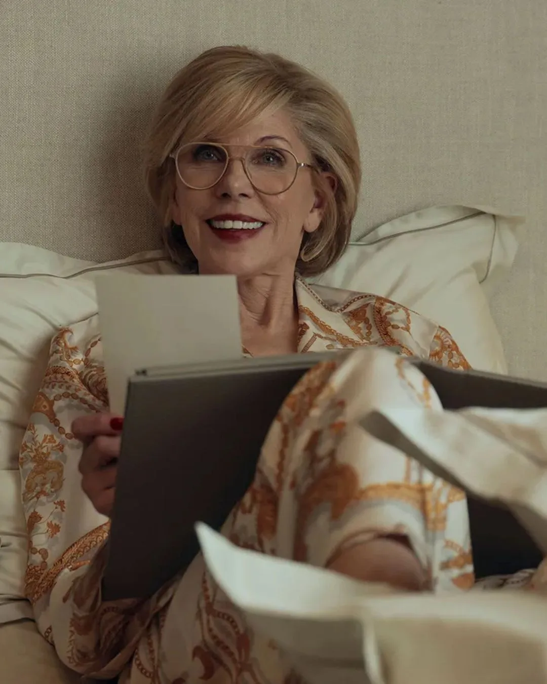 A woman with short blonde hair and glasses smiling as she lies in bed wearing silk pyjamas, reading a book. 