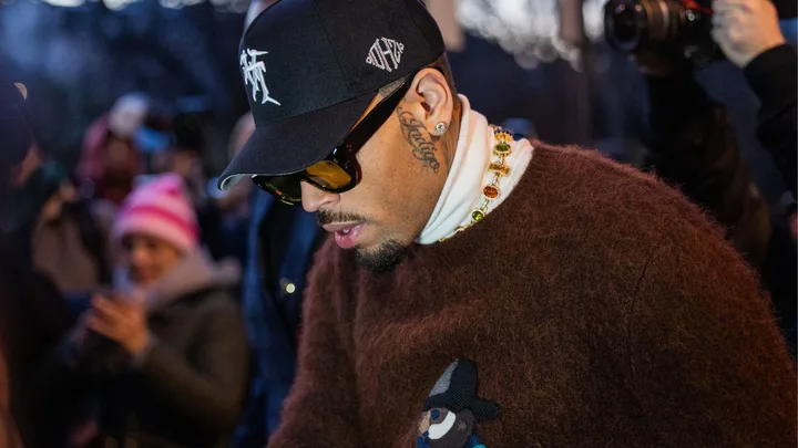 Rapper Chris Brown wearing a baseball cap and sunglasses and a brown jumper looking down in a crowd of people.