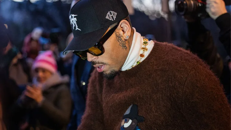 Rapper Chris Brown wearing a baseball cap and sunglasses and a brown jumper looking down in a crowd of people.