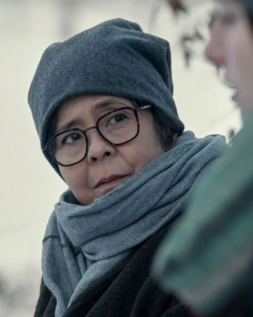 A woman wearing large glasses, a grey beanie and large grey scarf looking serious.