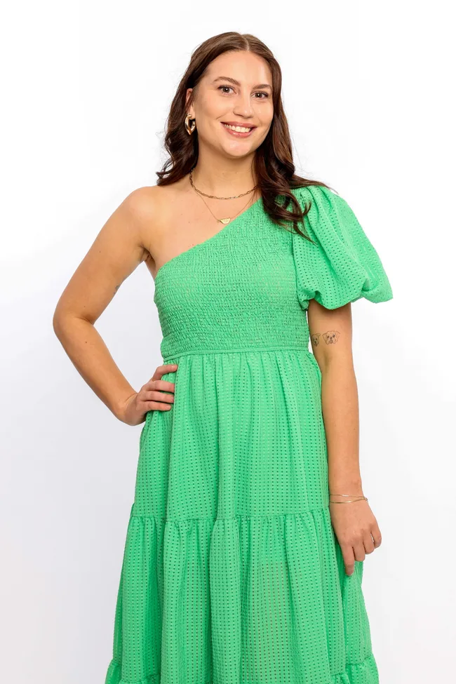 A brunette woman stands with her hand on her hip wearing a green off the shoulder dress.