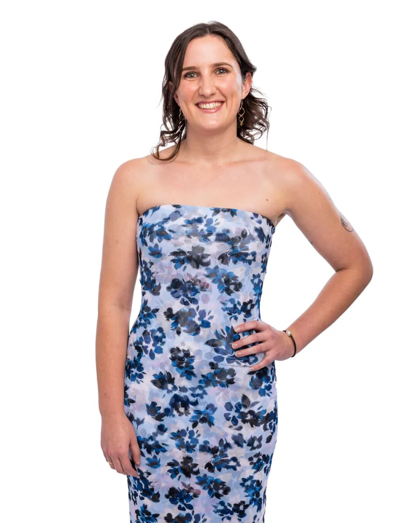 A brunette woman wearing a blue floral strapless dress standing with one hand on her hip.