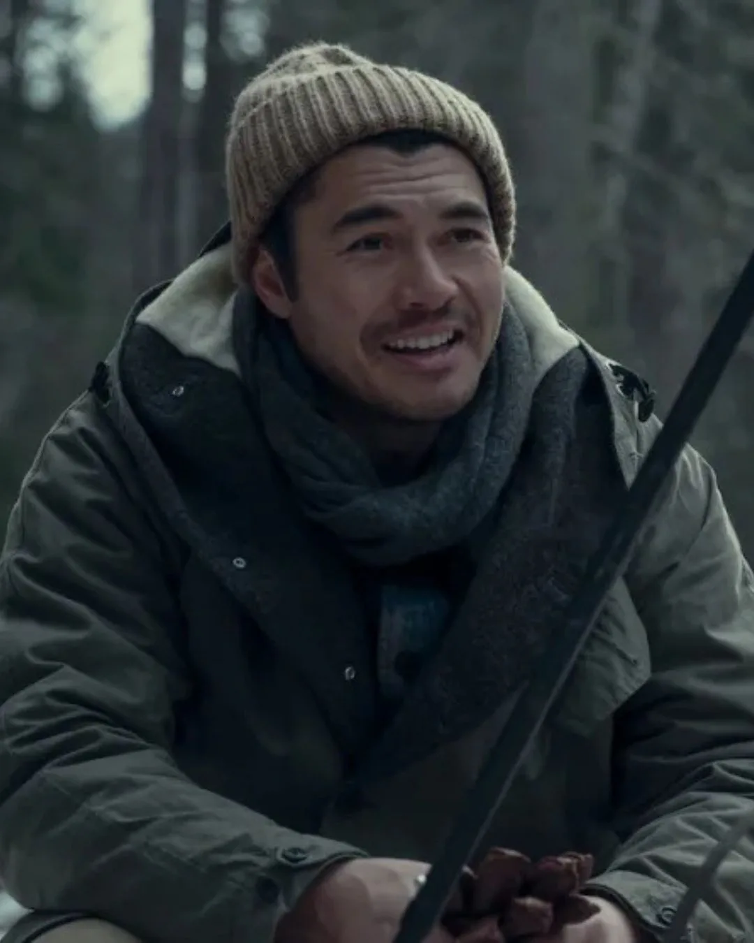 Actor Henry Golding on the set of Nine Perfect strangers. He's wearing a taupe coloured beanie and khaki ski gear and is sitting at a campside fire. 