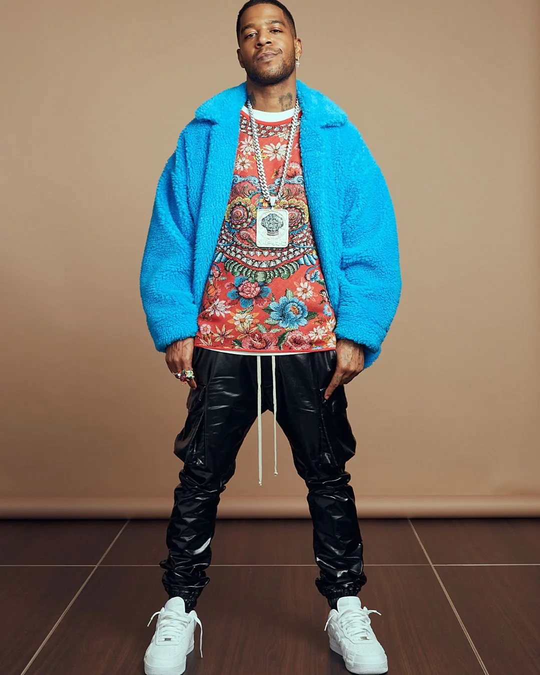 A man wearing a blue jacket and colourful t-shirt with black leather pants and white sneakers standing with his legs apart looking at the camera. 