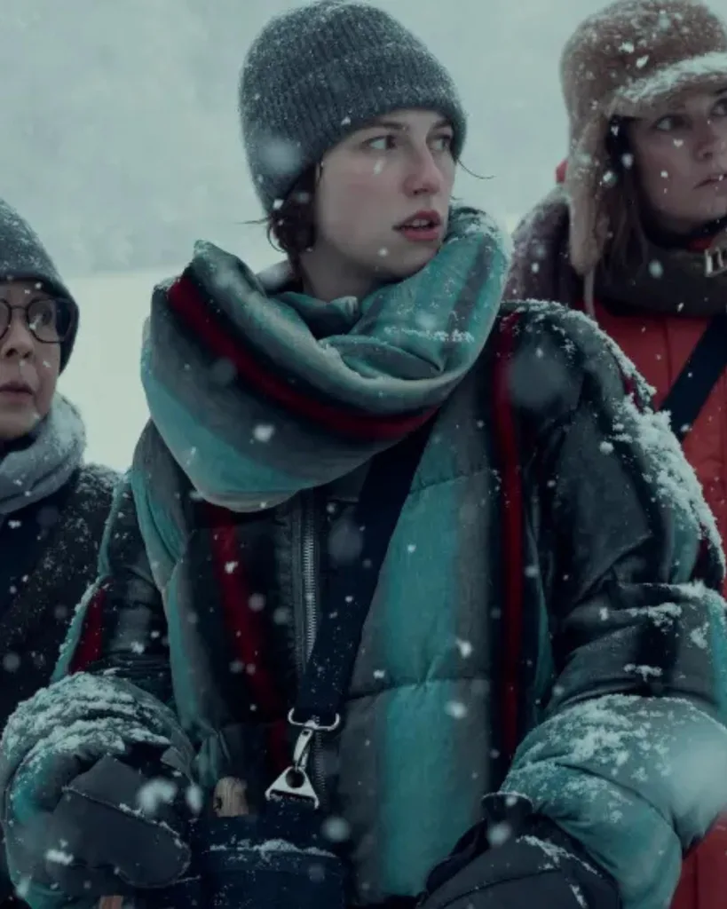 A woman wearing a beanie, scarf and grey/green snow gear standing in the snow.