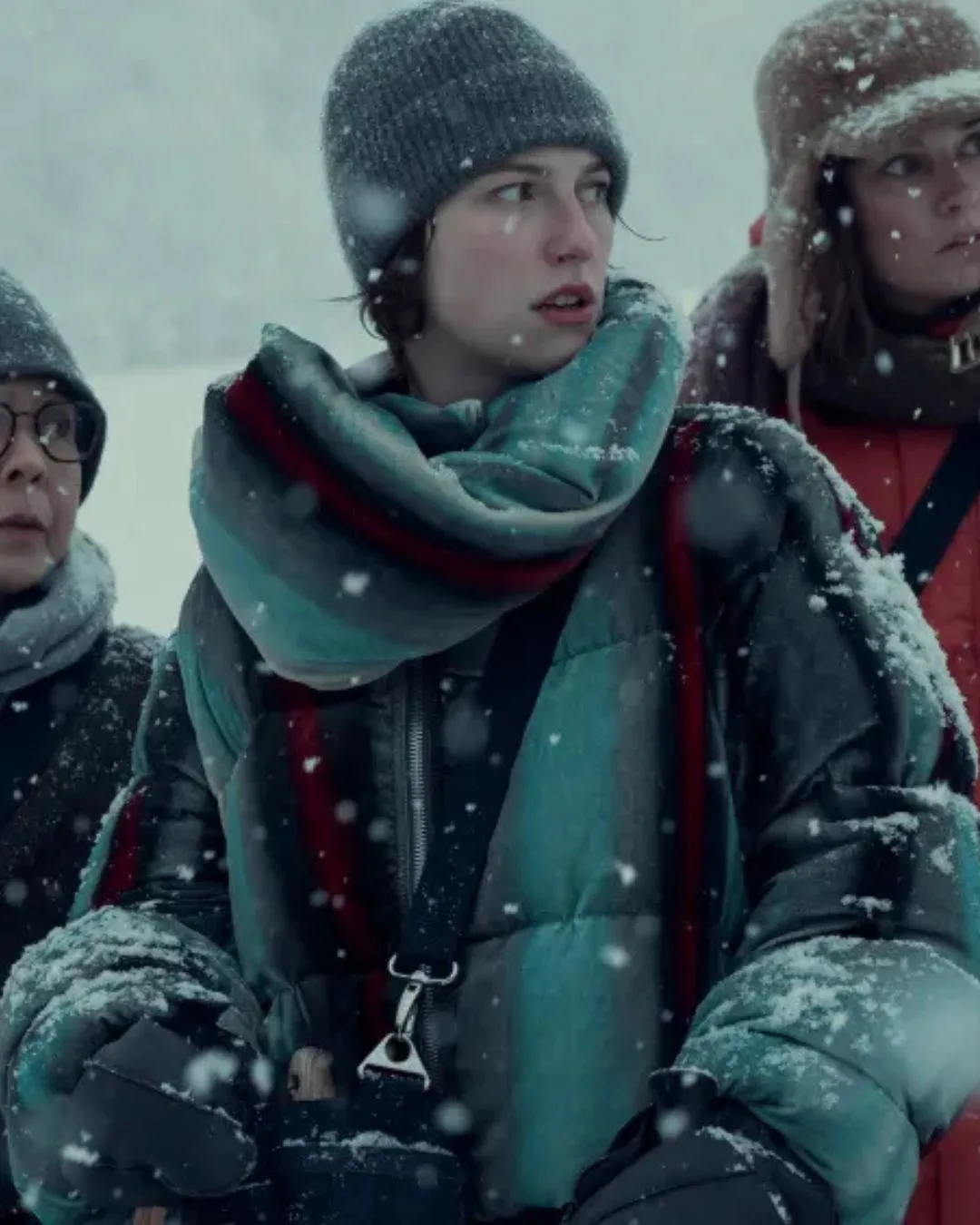 A woman wearing a beanie, scarf and grey/green snow gear standing in the snow. 
