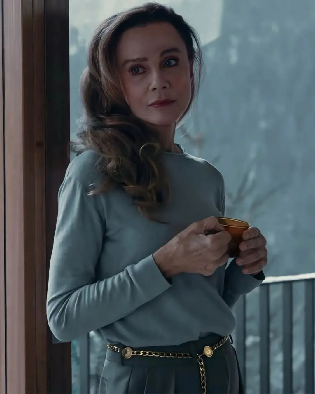 A woman with long brunette hair leaning against a window holding a tea cup. She wears a grey  long sleeve top and grey suit pants. 