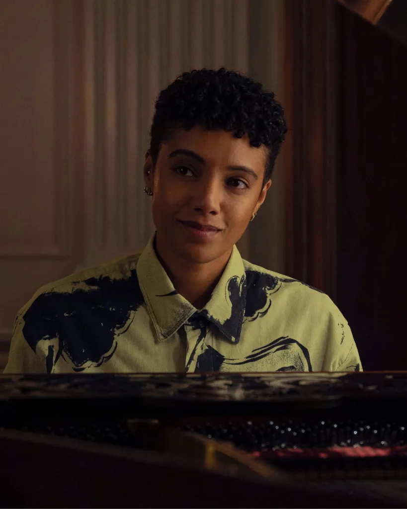 A person with short cropped hair and a button up white and blue shirt sitting at a piano.