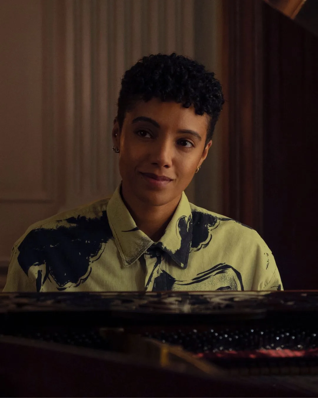 A person with short cropped hair and a button up white and blue shirt sitting at a piano. 