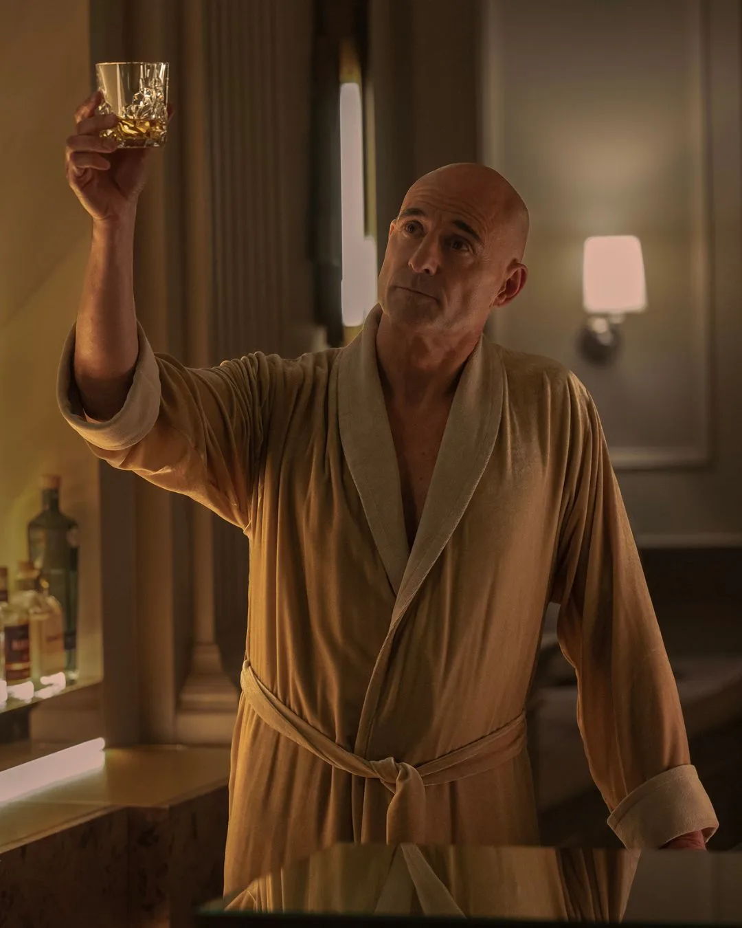 Actor Mark Strong - a bald man standing behind a bar dressed in a camel-coloured robe holding up a glass of whiskey. 