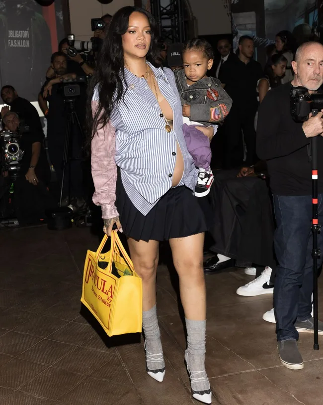 A woman wearing a striped shirt and black mini skirt carrying her child on her hip.