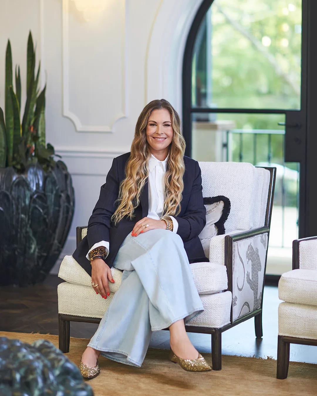 A woman with long blonde hair sitting on a chair wearing light blue pants and a navy blazer. 