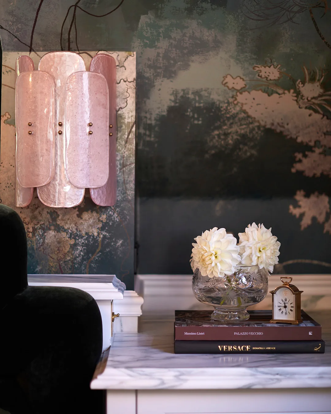 A close up shot of a bedside table with green wallpaper on the back wall and a pink scalloped Murano glass wall light. 