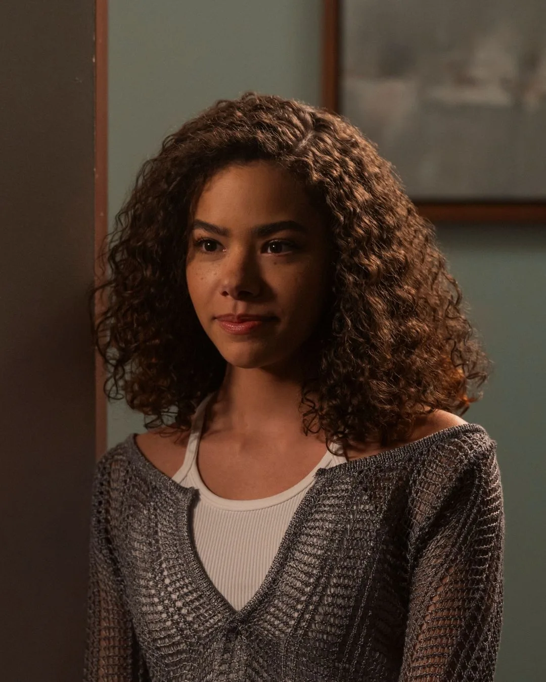 A young woman with curly hair wearing a grey cardigan, smiling.