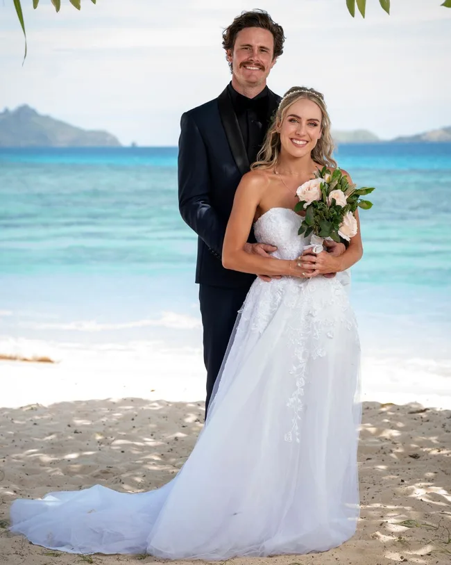 A man and woman dressed in wedding attire standing and smiling on a beach.