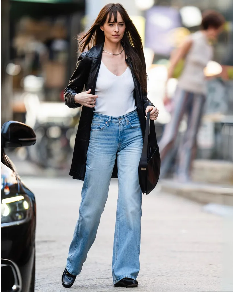A woman walking wearing blue jeans a white tank top and long black leather jacket.