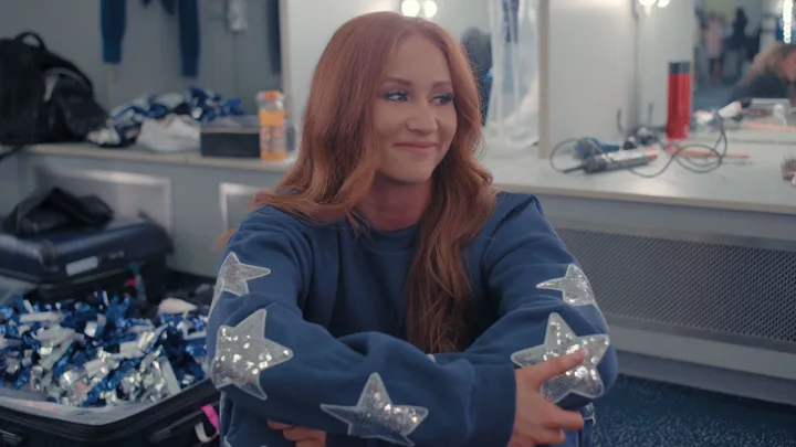 A red-haired woman sitting and smiling in a dressing room wearing a blue jumper with sequin stars lining the arms.