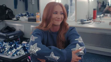 A red-haired woman sitting and smiling in a dressing room wearing a blue jumper with sequin stars lining the arms.