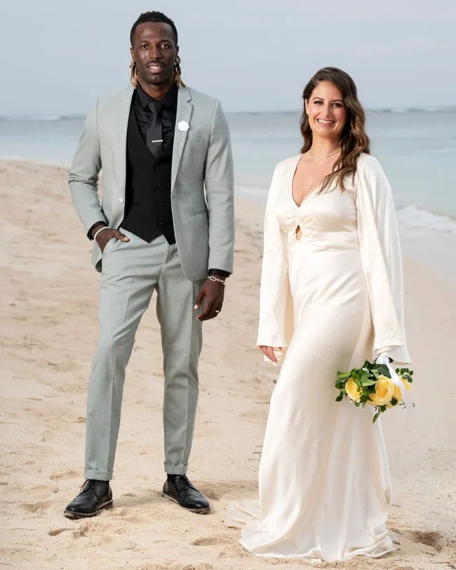 A man and woman dressed in wedding attire standing and smiling on a beach.