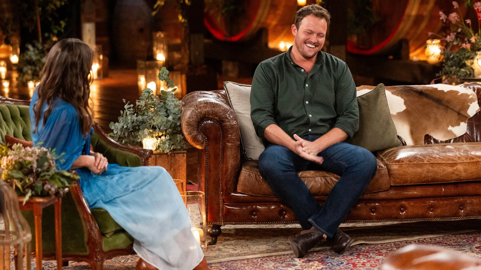 A smiling man wearing a green shirt and jeans talks to a woman wearing a long blue dress on a lounge. 