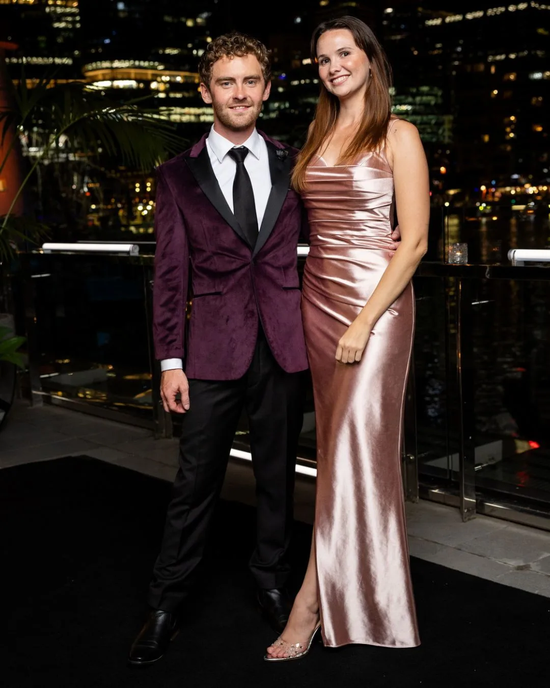 A man wearing a burgundy suit standing next to a young woman in a cream silk gown. 