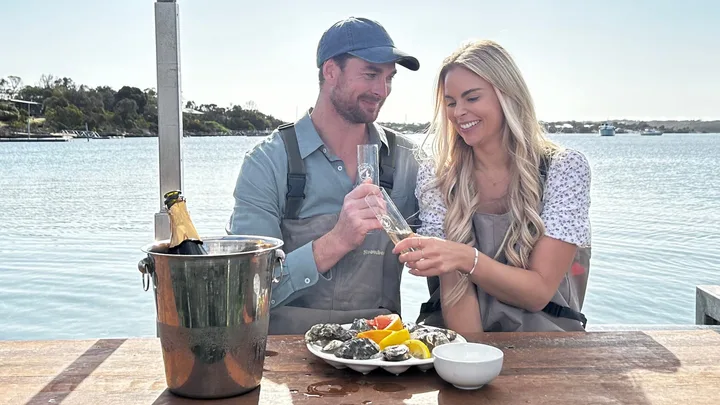 A man and a woman toasting with a glass a champagne while shucking oysters.