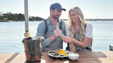 A man and a woman toasting with a glass a champagne while shucking oysters.