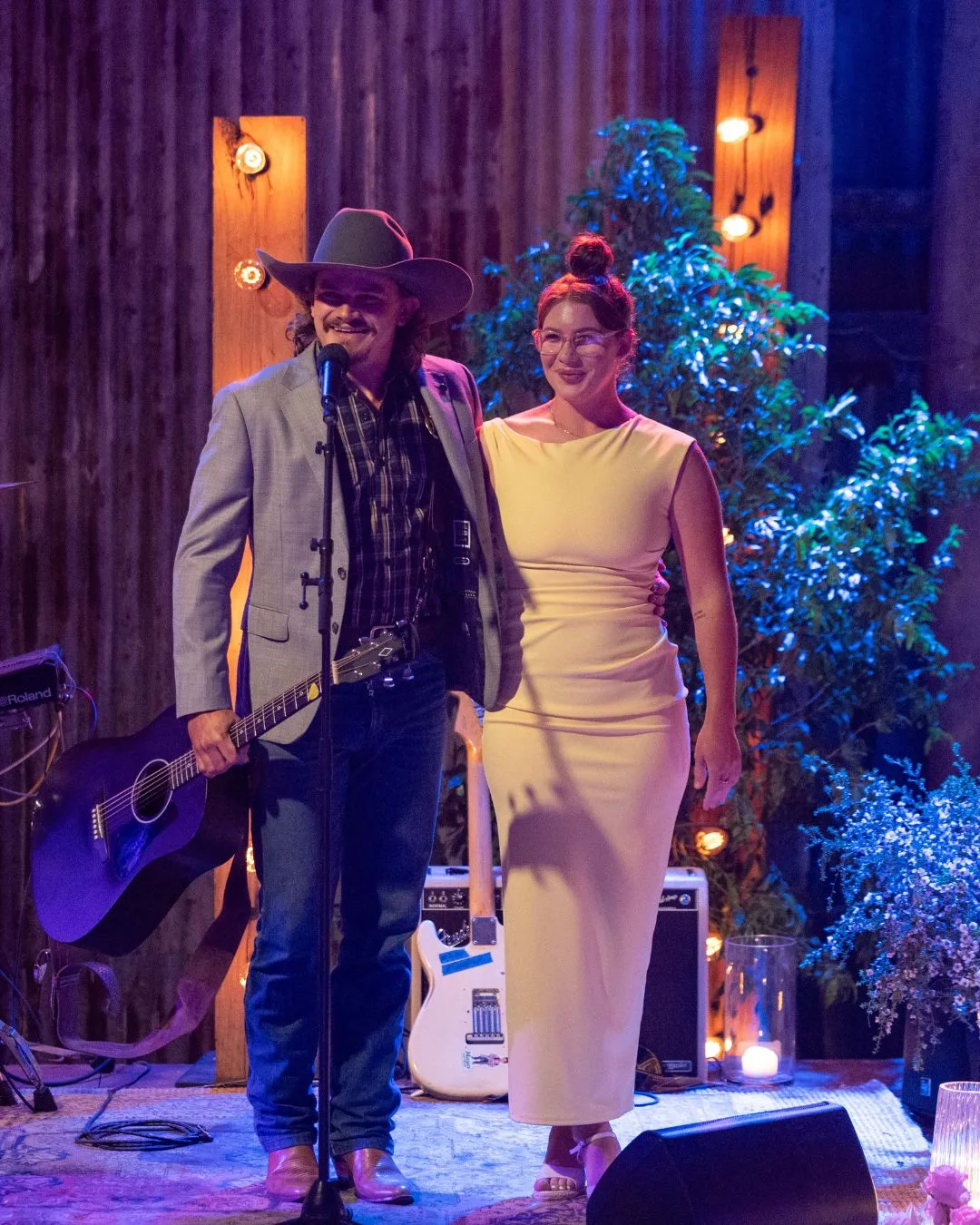 A man wearing a cowboy hat, holding a guitar, stands on stage ready to perform with a woman wearing a white dress by his side. 