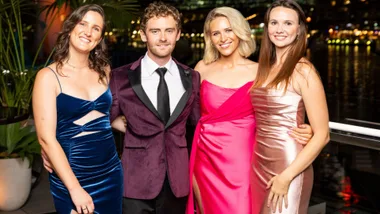 A man wearing a burgundy suit standing with three women wearing evening dresses, smiling.
