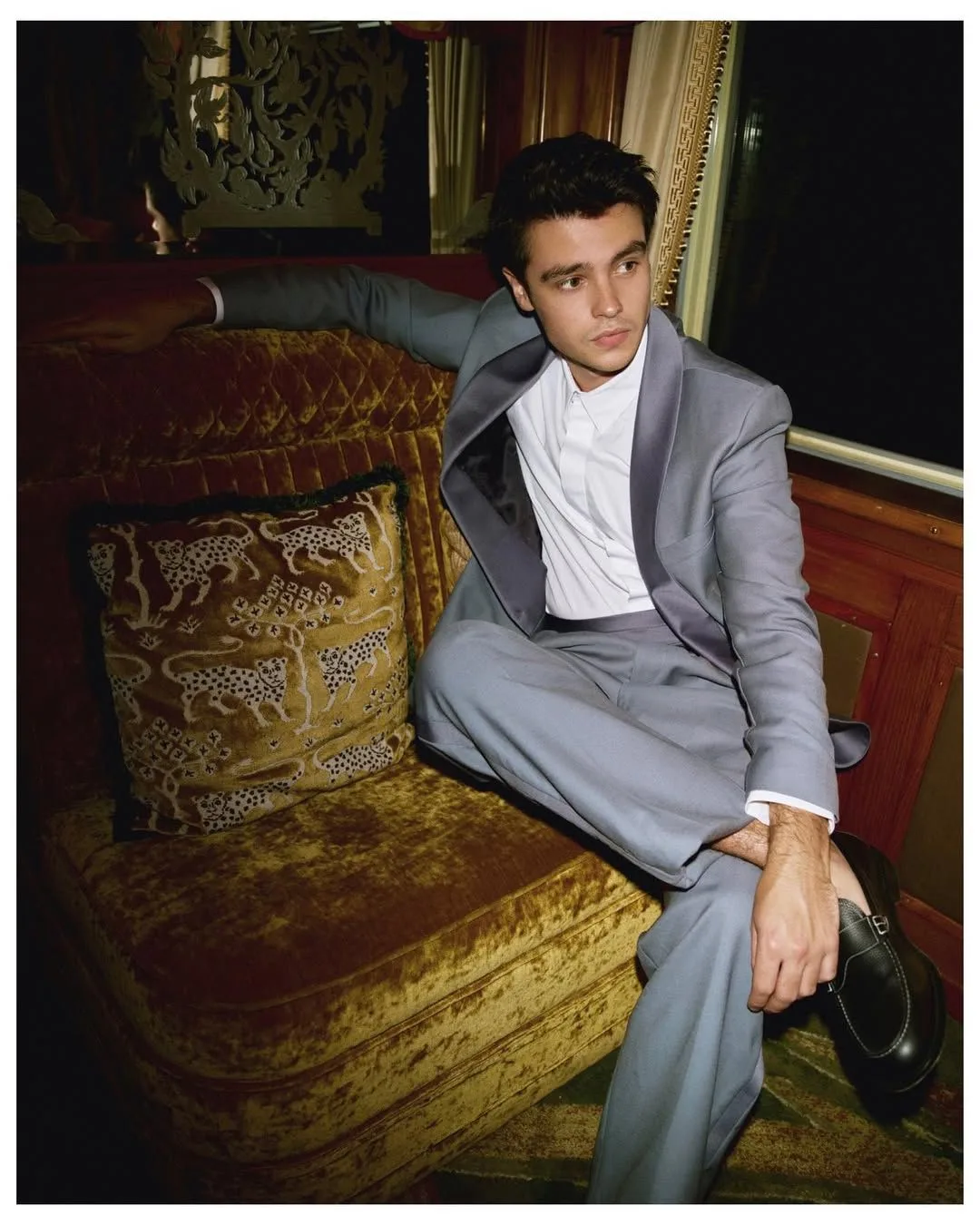 A man sitting on a velvet sofa wearing a light grey suit. 