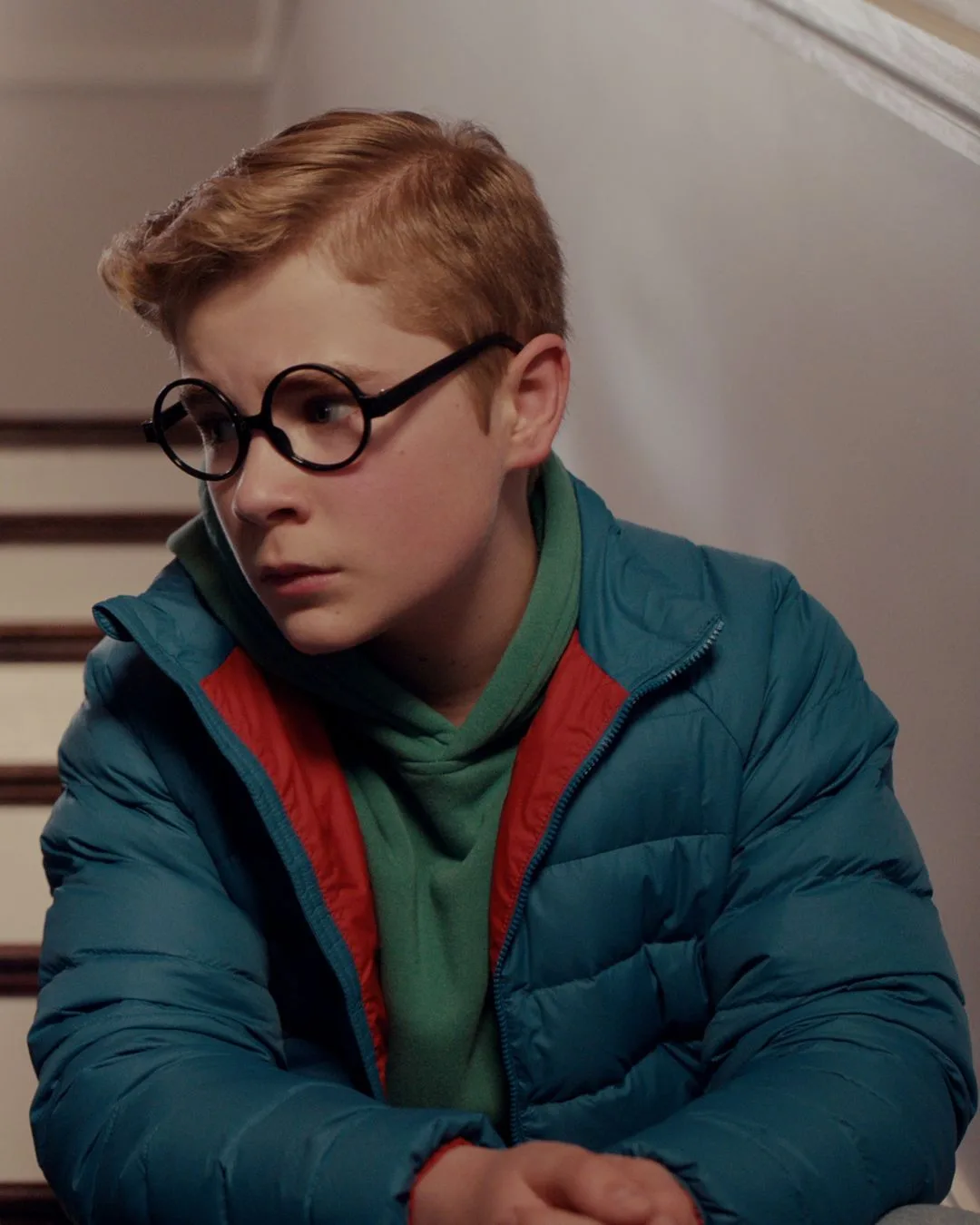 A young blonde boy in a blue puffer jacket with round black framed glasses looking concerned.