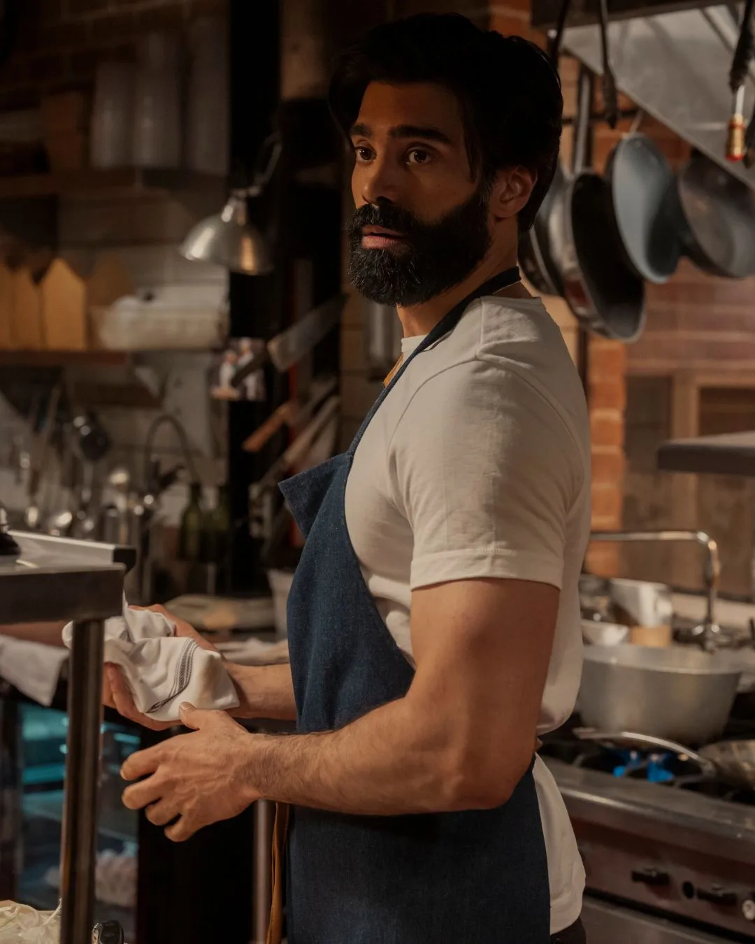 A bearded man wearing a white t shirt and blue apron in the kitchen.
