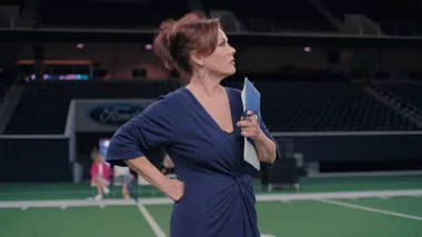 A red-haired woman standing in the middle of a NFL field holding a clipboard.