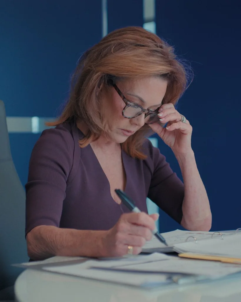 A red haired woman wearing glasses writing on a table.