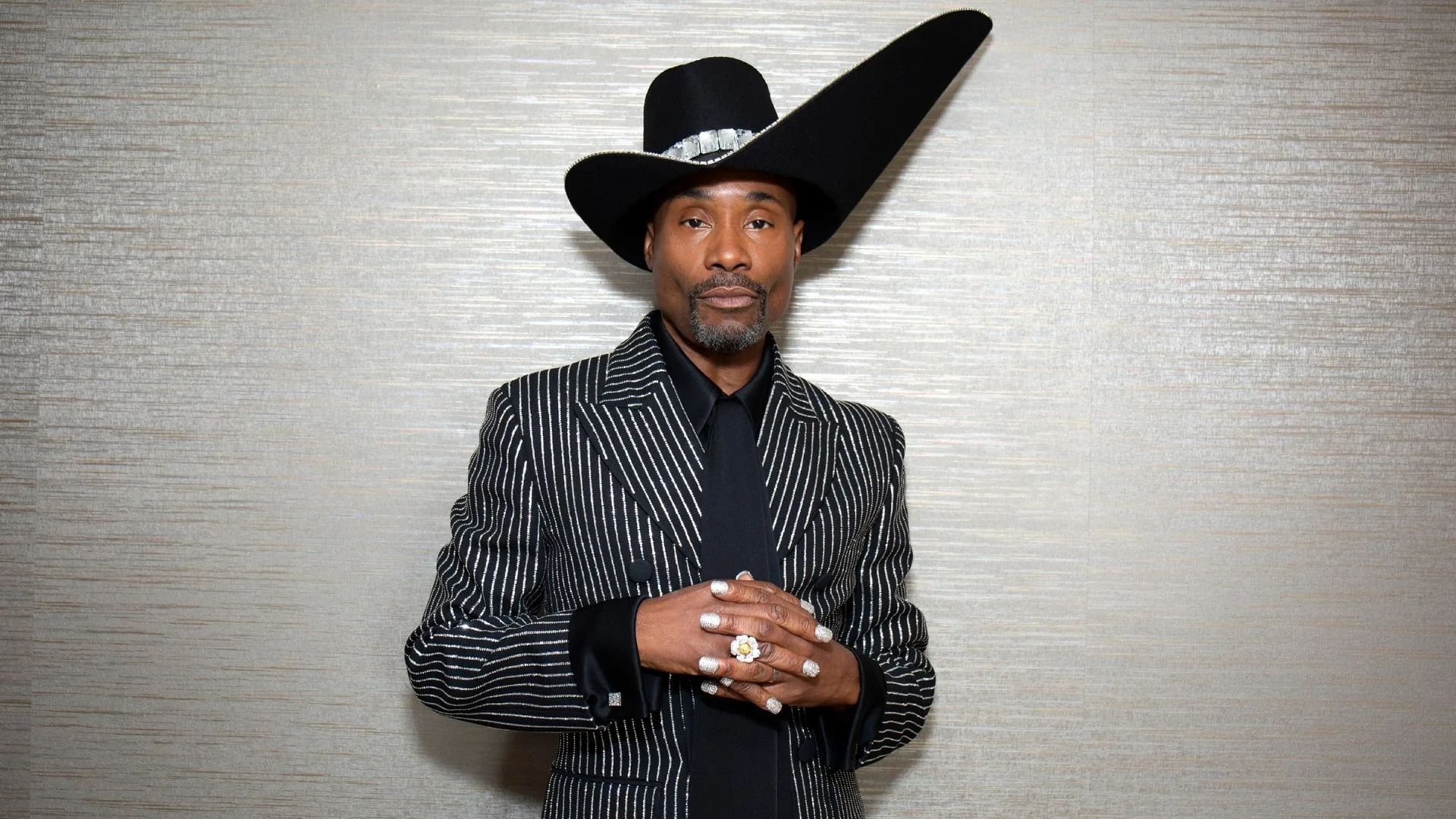A man wearing a statement hat clasping his hands together. 