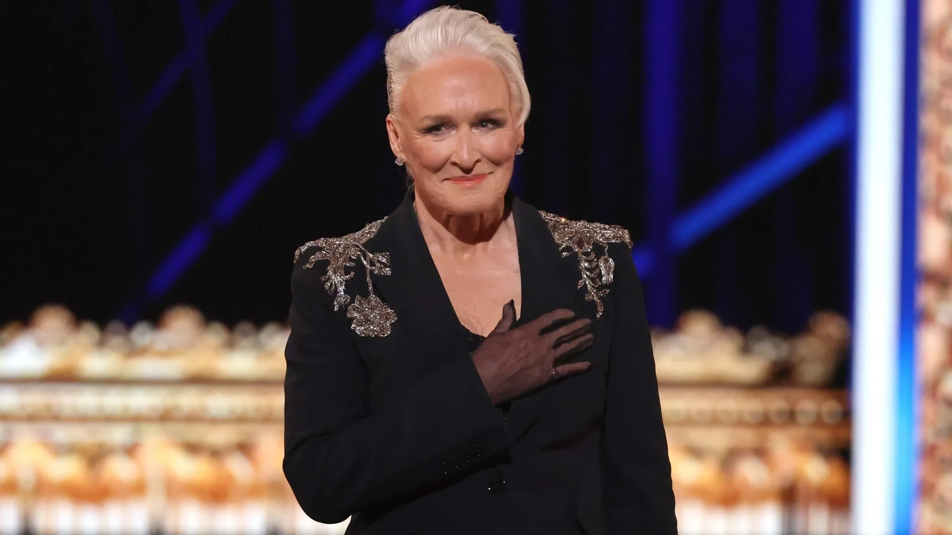 A woman with short grey hair wearing an embellished black blazer holds her hand across her heart. 