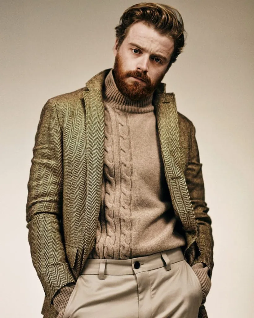 A man with red hair and red beard wearing a tan jumper, green herringbone blazer and cream pants standing with his hands in his pockets.