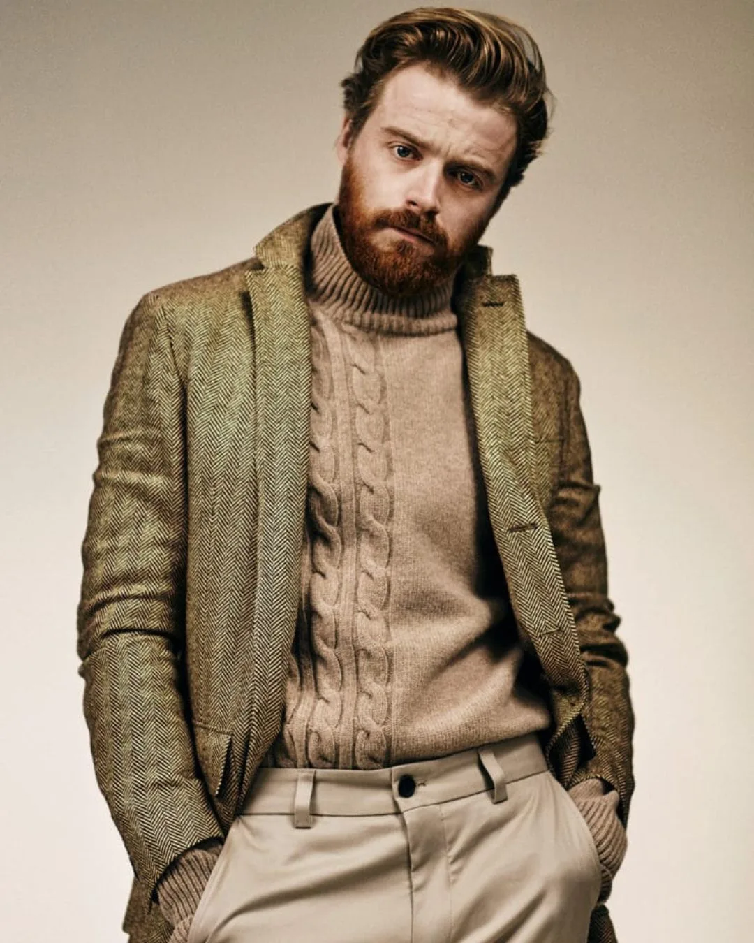 A man with red hair and red beard wearing a tan jumper, green herringbone blazer and cream pants standing with his hands in his pockets.