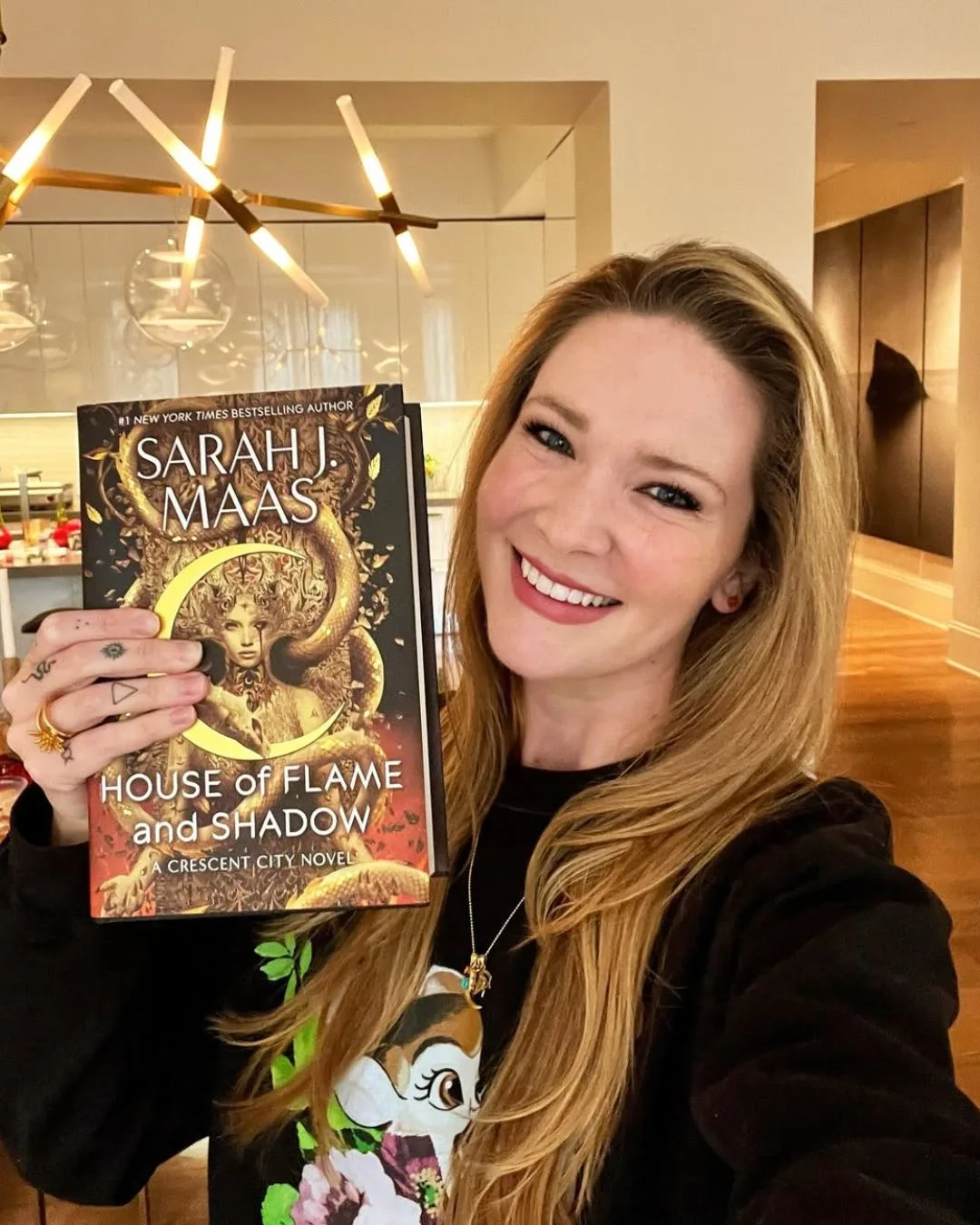 A blonde woman smiling while holding up a book. 