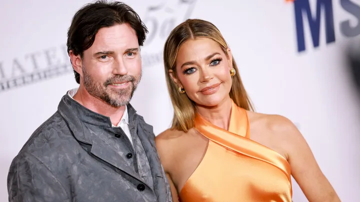 Denise Richards and her husband pose smiling. A woman wearing a light orange dress and a man wearing a grey blazer and white shirt smiling.