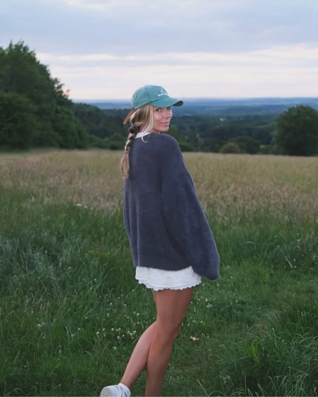 A girl standing in a field wearing a  baseball cap and navy jumper. 