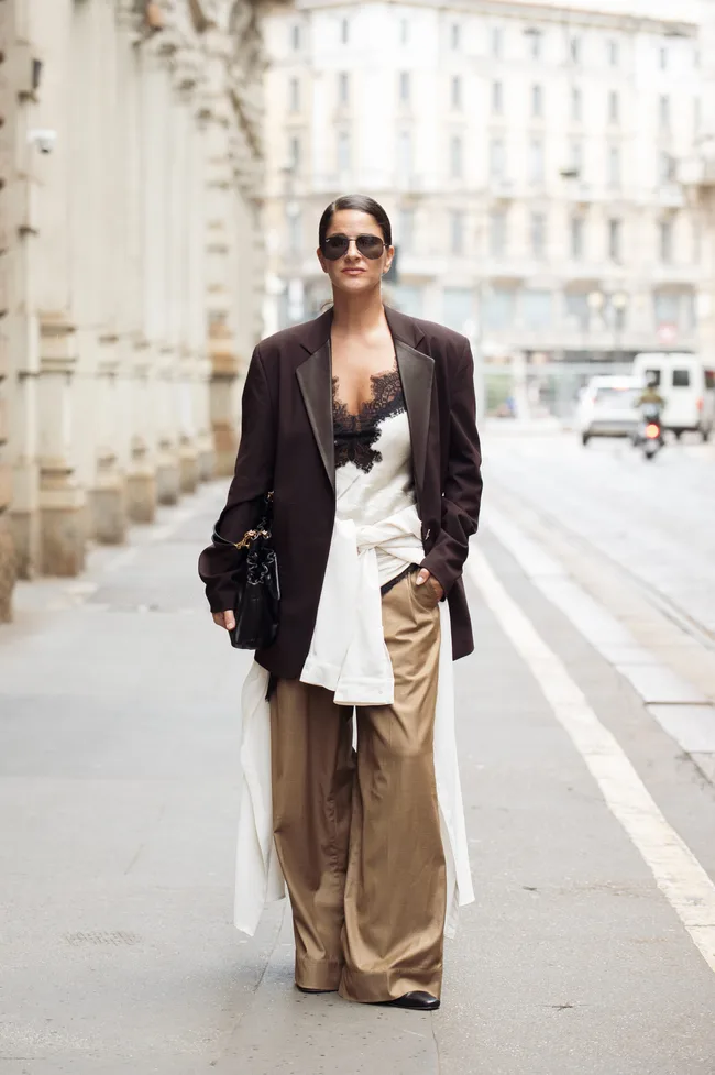 Gili Biegun wears plum brown blazer, beige shirt tied around the waist, light brown trousers and white silk asymmetrical dress
