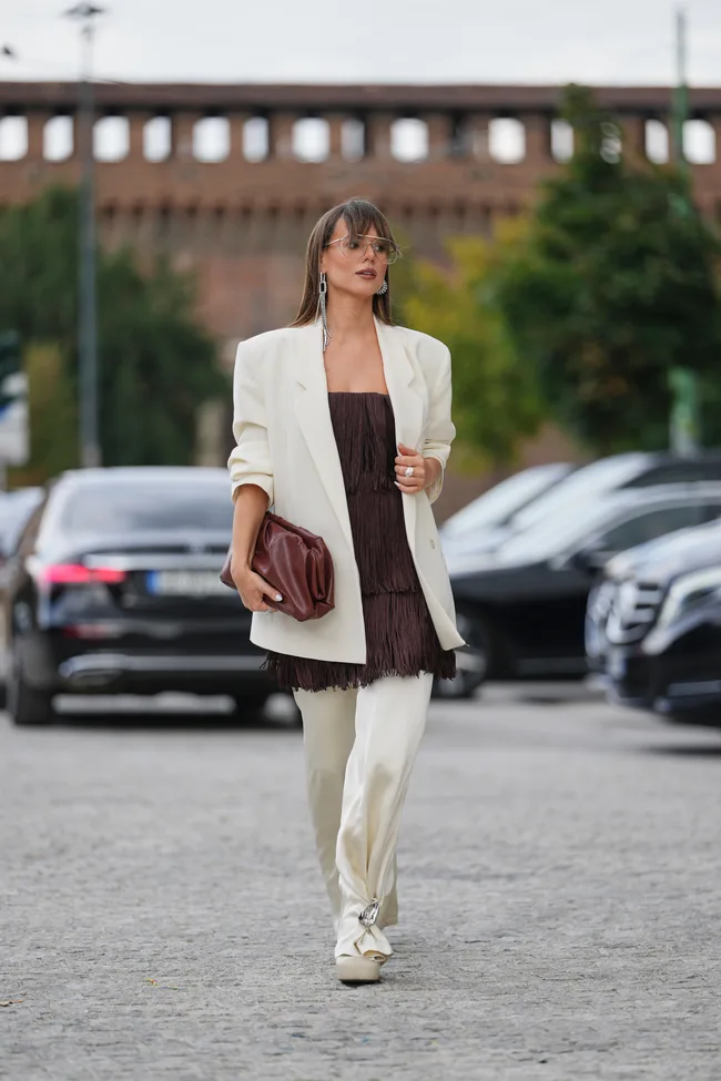 Milan Fashion Week guest wears long straight brown hair with blunt bangs and a center part, clear oversized glasses, silver-tone crystal drop earrings and silver rings, a brown leather clutch bag, a cream tailored oversized blazer jacket over a strapless brown fringed slip dress with fringe trim and matching cream wide-leg trousers