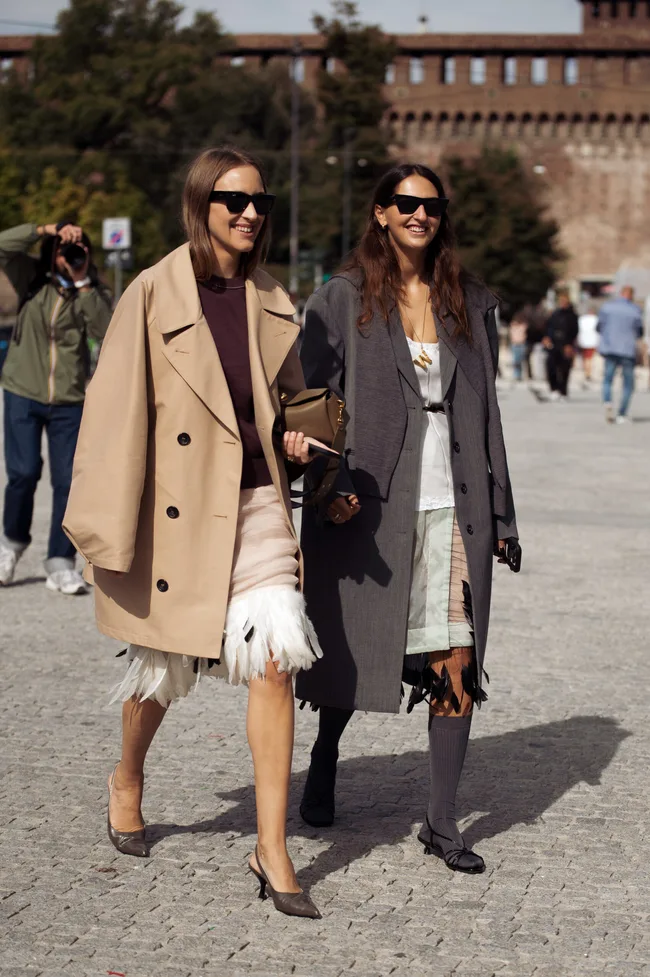 Michela D'Angelo wears a beige trench coat over a dark top and a nude skirt with white feather trim paired with pointed metallic heels and a taupe clutch bag a