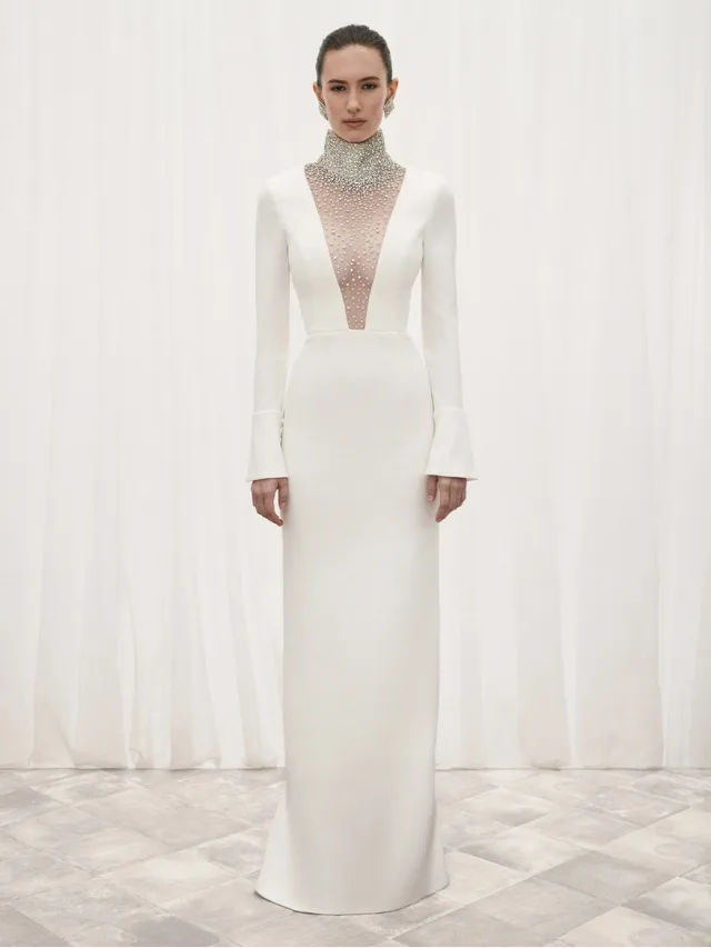 woman stands still for a portrait wearing a long-sleeve rebecca vallance white wedding gown with silver detail over a plunging neckline