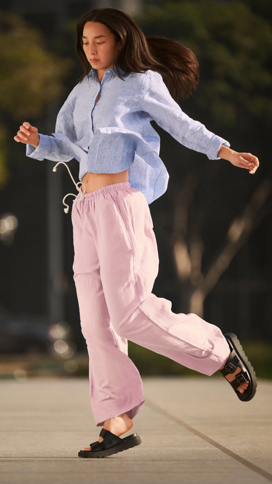 A model with long hair wearing a blue and white striped shirt and pink pants skipping outside.