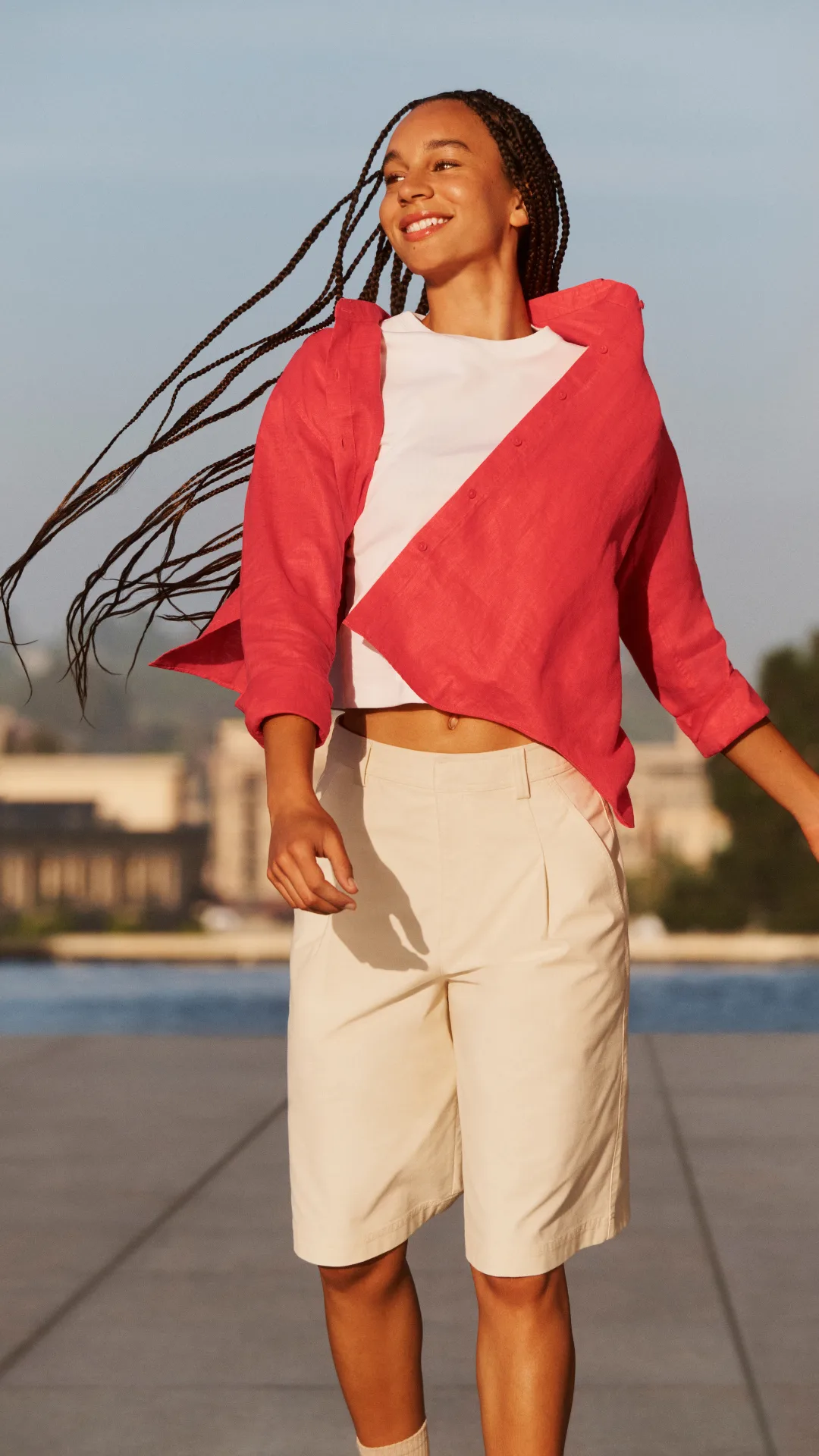 A model with long braids wearing a red shirt and beige shorts skipping outside.