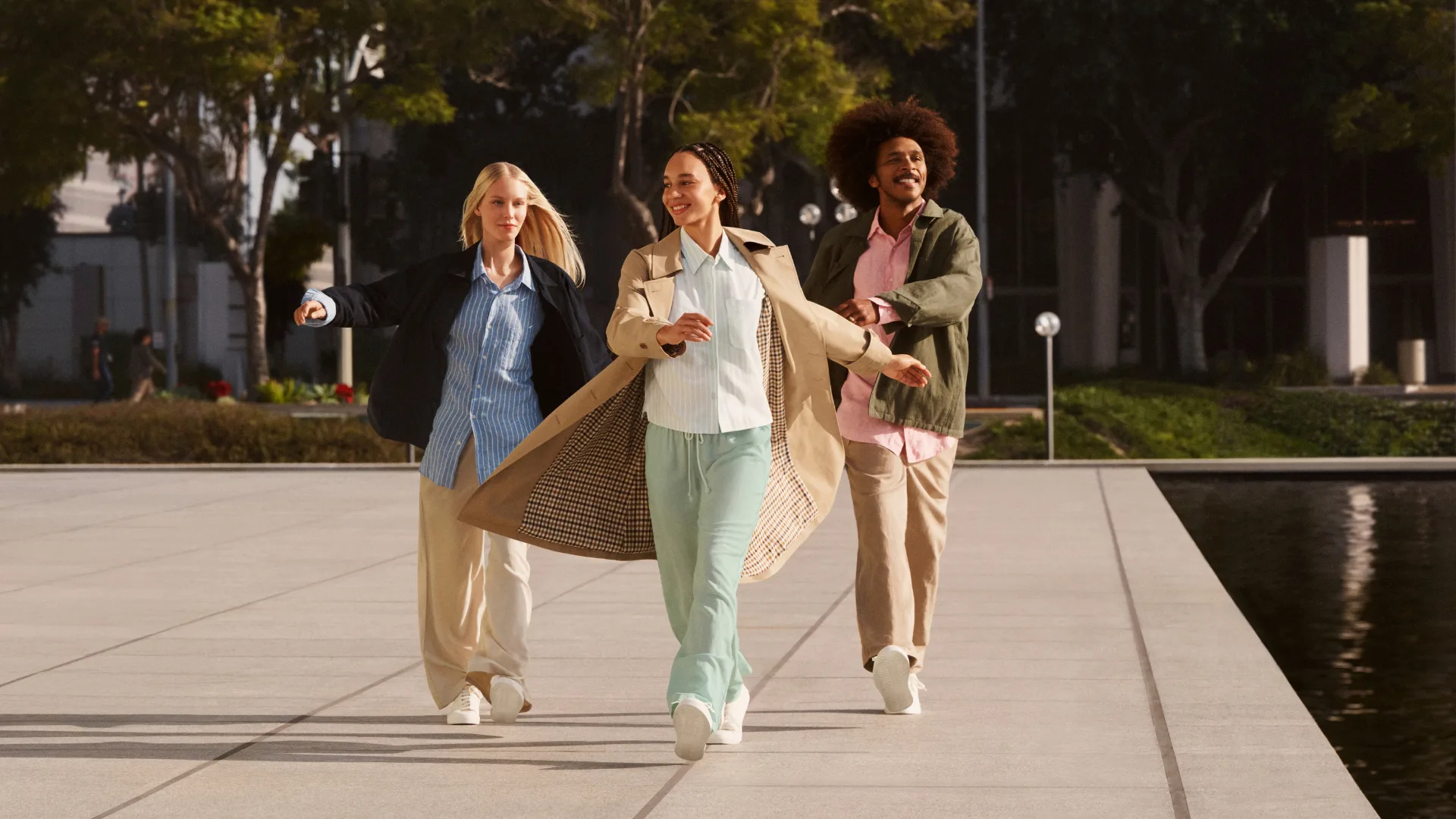 Three models: one blonde woman wearing a blue and white stripe shirt and beige pants; a dark-haired woman wearing a striped shirt and green pants; and a man wearing a pink shirt and beige pants skipping outside