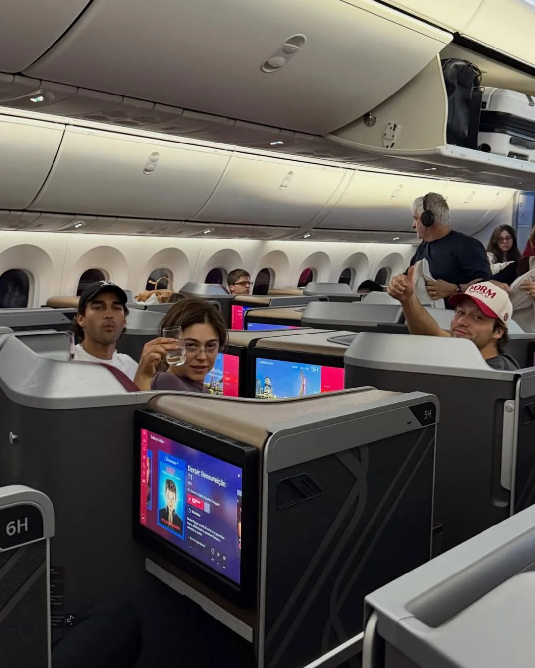 mika abdalla and josh heuston sit in business class on a flight while holding up glasses of champagne
